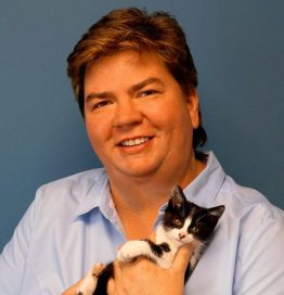 Bio portrait of Alice Burton, smiling in a pale blue shirt against a dark blue background, holding a black and white kitten